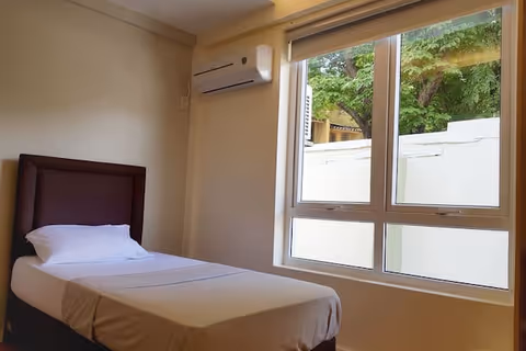 A simple bedroom with a single bed made with white sheets and a pillow, a brown padded headboard, a large window with a view of trees and a white wall outside, and an air conditioning unit mounted on the wall above the bed.