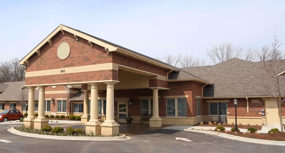 Exterior view of a single-story brick building with a covered entrance supported by large columns, surrounded by a parking area and some landscaping with small bushes and trees.