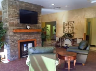 A cozy senior living facility common area with a stone fireplace, a mounted flat-screen TV above the mantel, a clock on the mantel, two green leather armchairs, a round wooden coffee table, a decorative wall piece, and plants in the background.