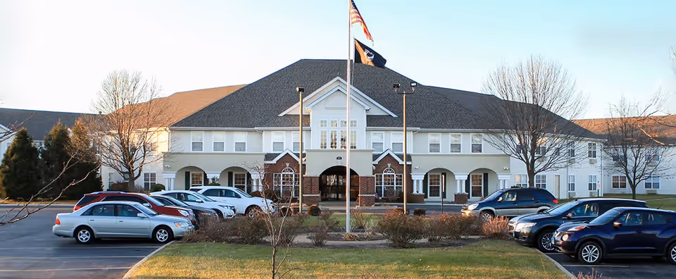Front exterior view of The Pointe at Jacksonville senior living facility with a parking lot in front, several parked cars, a flagpole with American and other flags, and a two-story building with white and brick facade under a clear sky.
