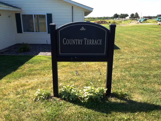 A black sign with white lettering that reads 'Country Terrace' standing on a grassy lawn in front of a white building with a window and black shutters.