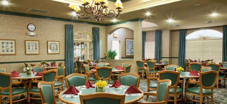 A dining room in a senior living facility with multiple round tables set with green chairs, red folded napkins, silverware, and small flower arrangements. The room has beige walls with framed artwork, green curtains, a chandelier, and a clock on the wall.