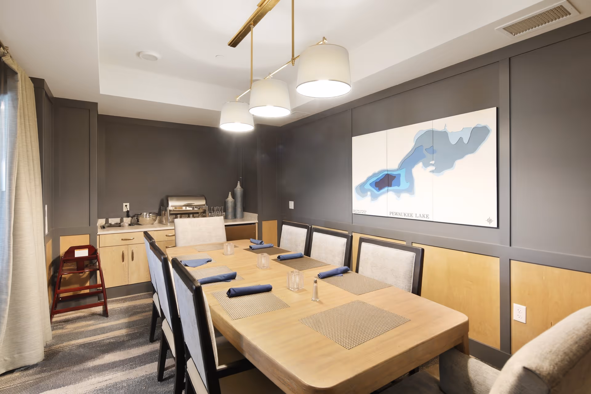 A dining room with a wooden table set for six people with placemats and folded blue napkins. The room has gray walls with light wood paneling on the lower half and a large map of Pewaukee Lake hanging on the wall. There are three pendant lights hanging above the table and a sideboard with decorative vases and serving dishes in the corner. A red high chair is placed near the sideboard.
