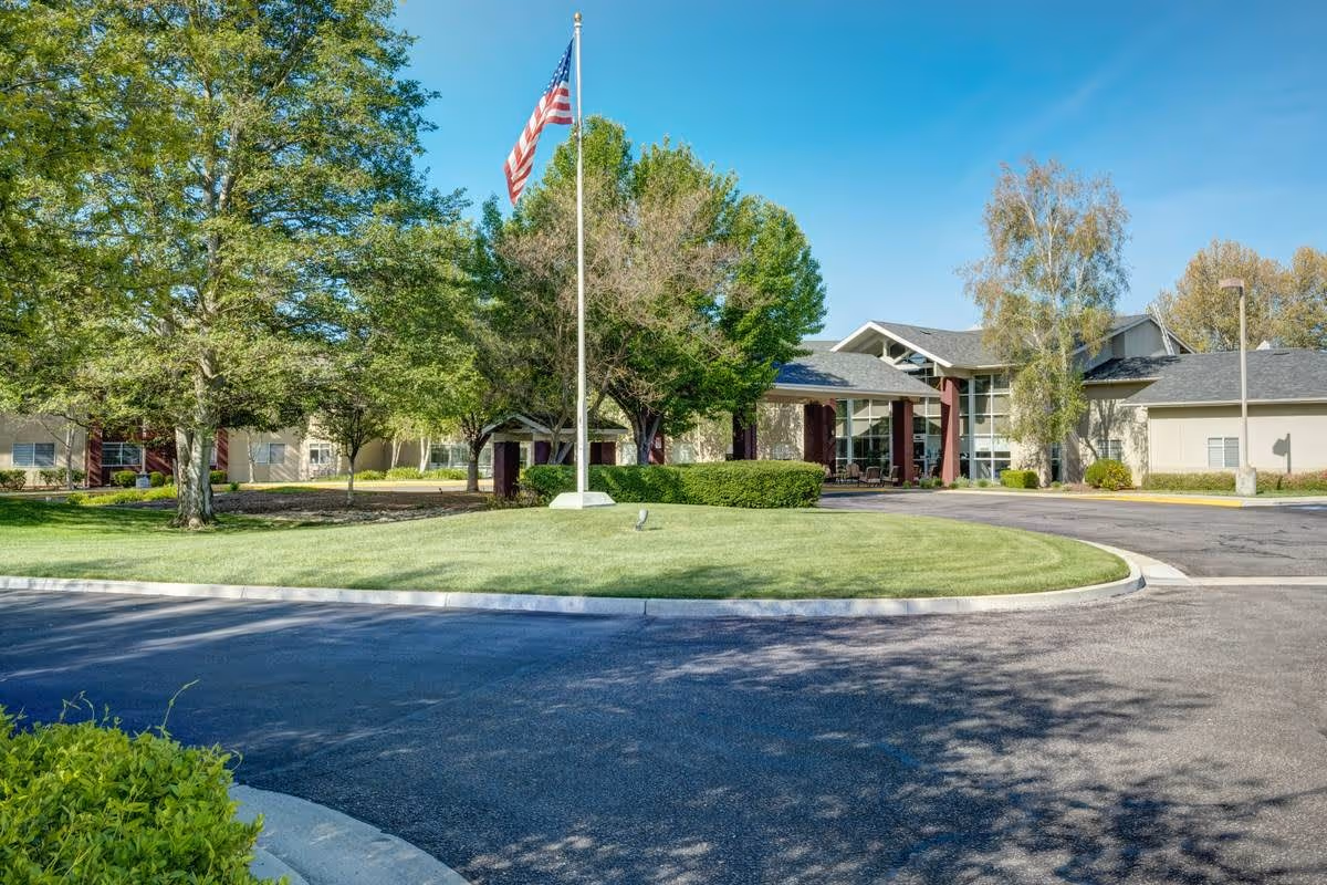 Exterior view of Creston Village senior living facility with a circular driveway, green lawn, trees, and an American flag on a flagpole under a clear blue sky.
