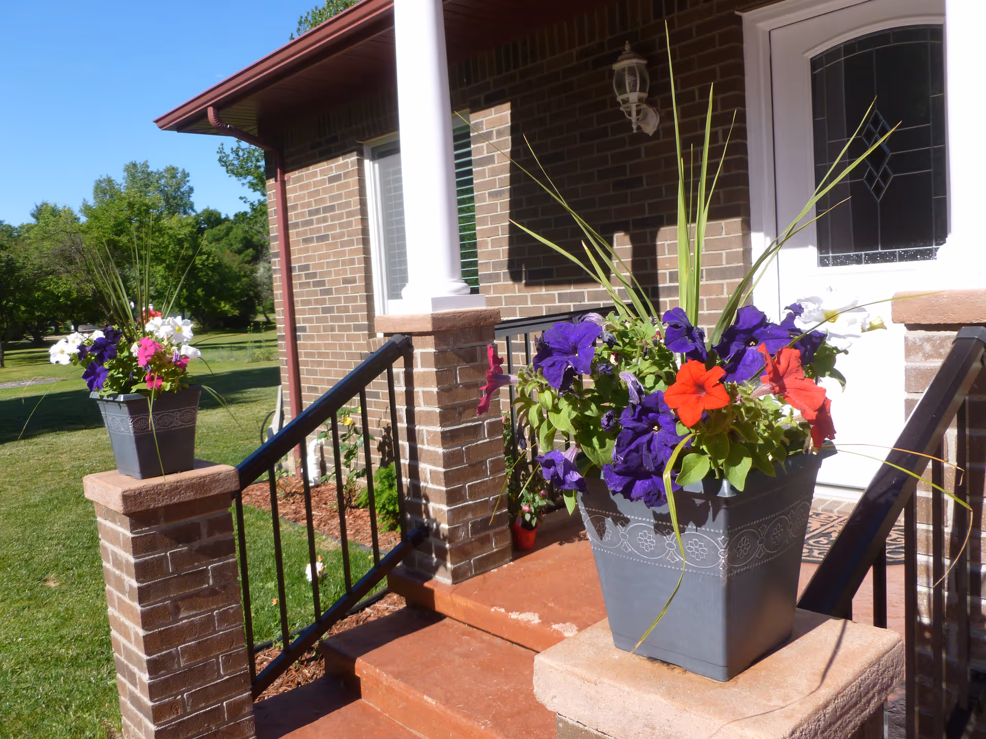 Front entrance of a brick building with steps leading up to a white door with decorative glass. Two large flower pots with colorful flowers, including purple and red blooms, are placed on brick pillars on either side of the steps. Green grass and trees are visible in the background under a clear blue sky.