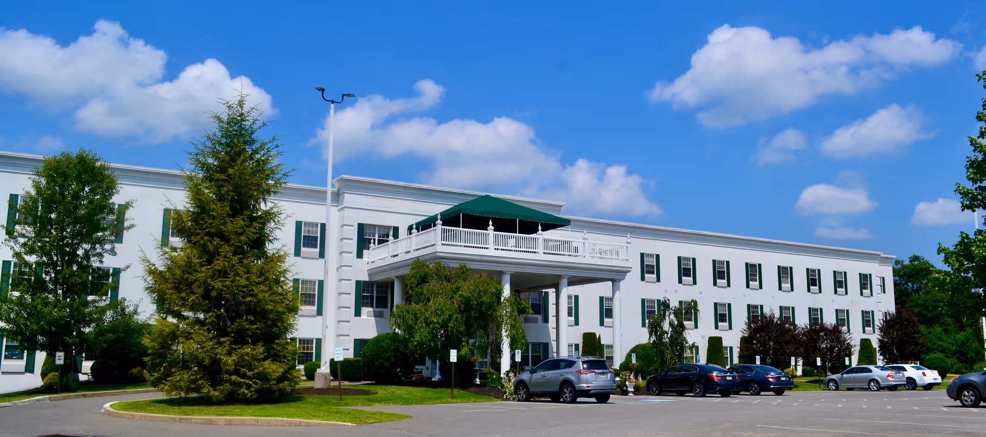 Exterior view of a large, white three-story senior living facility building with green window shutters and a covered balcony area. Several cars are parked in front of the building, and there are trees and shrubs around the parking lot under a blue sky with scattered clouds.