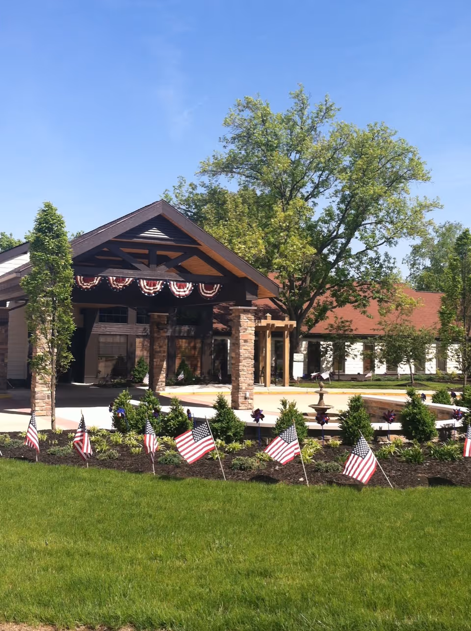 Exterior view of Riverview - An Optalis Health & Rehabilitation Center building entrance with a covered driveway supported by stone pillars. The area is decorated with American flags and patriotic bunting. There is a well-maintained lawn and trees under a clear blue sky.