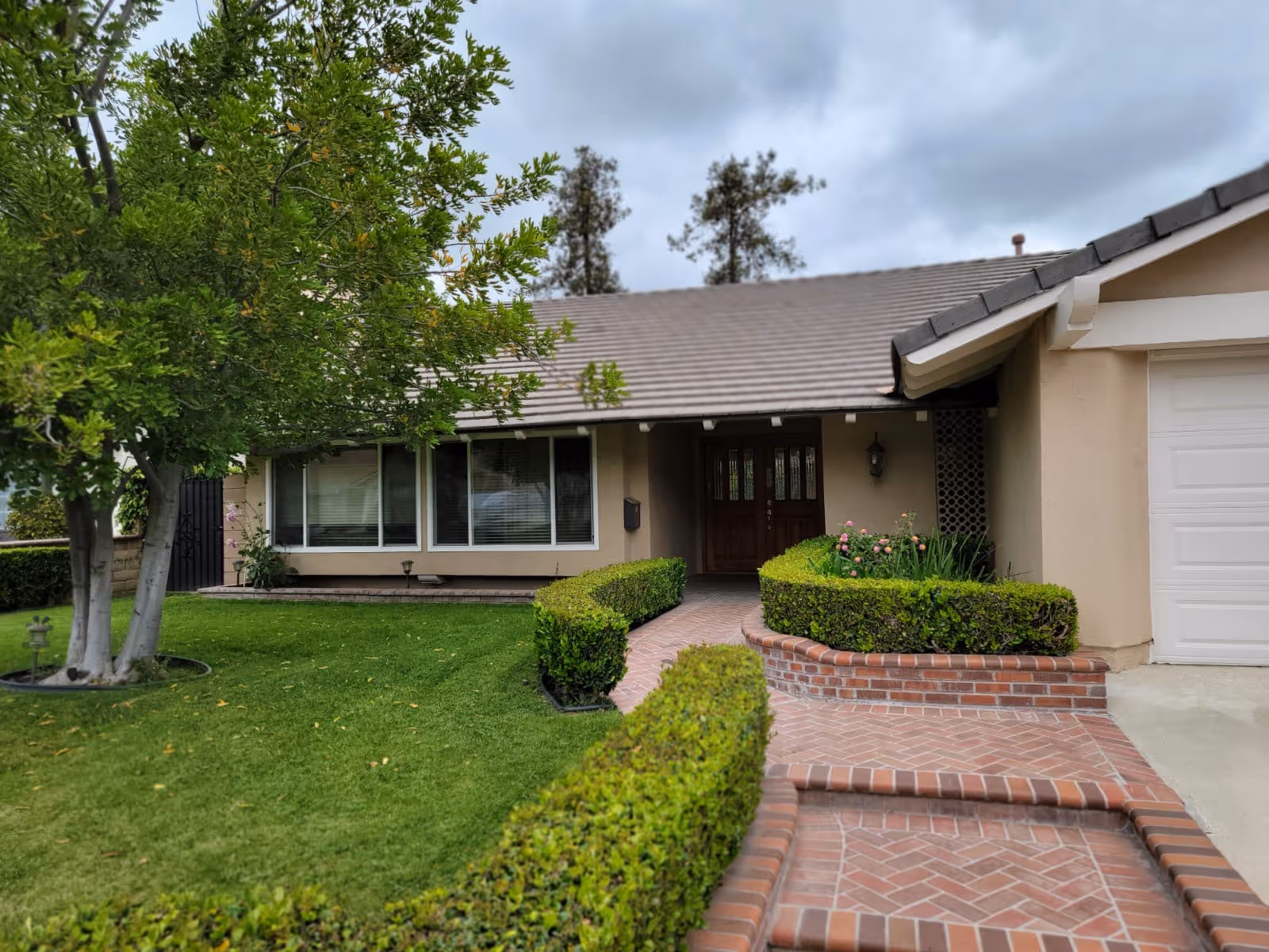 Front exterior view of a single-story house with a tiled roof, a brick walkway leading to a wooden front door, neatly trimmed hedges, a green lawn, and a tree on the left side under a cloudy sky.
