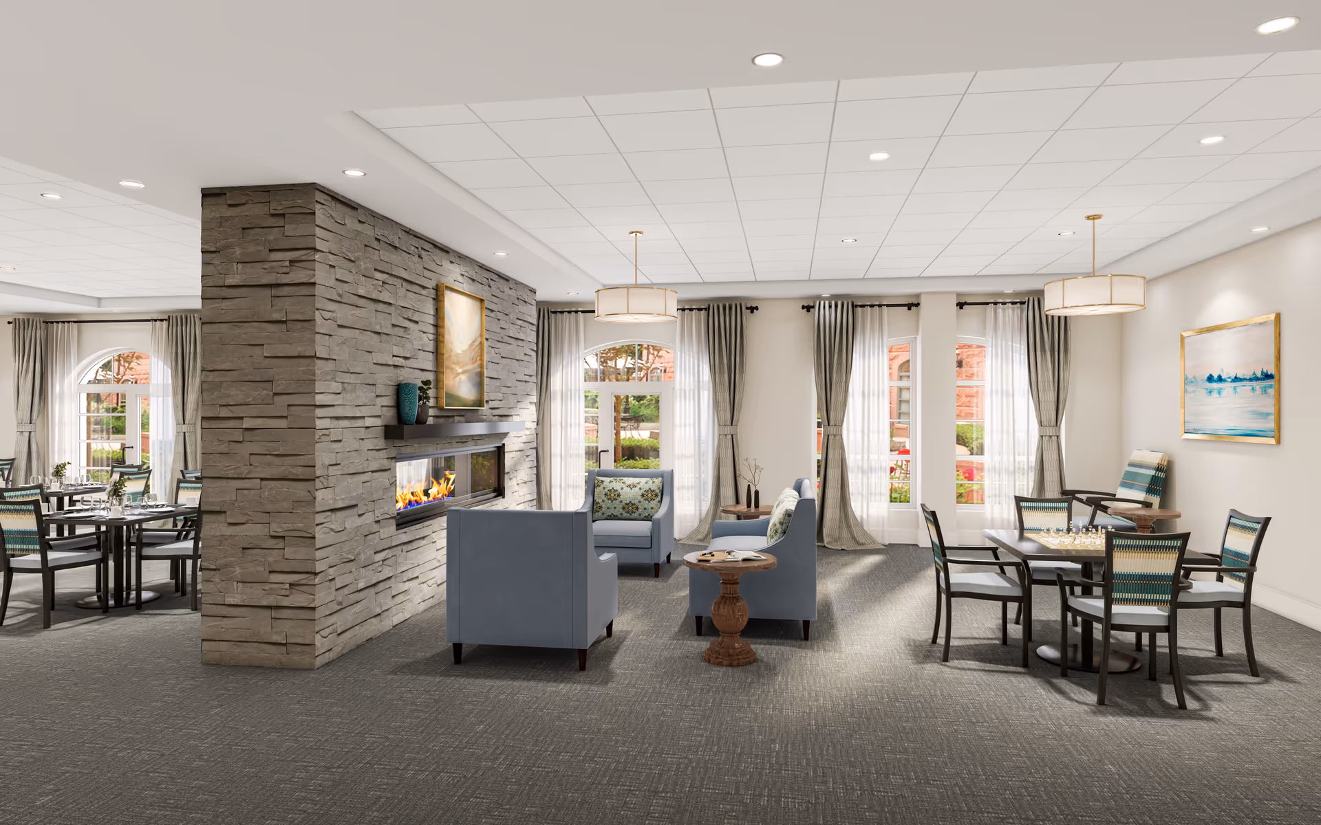 A spacious and well-lit common area in a senior living facility featuring a modern stone fireplace in the center. The room has multiple seating arrangements including blue armchairs around a small wooden table and dining tables with chairs. Large windows with light curtains allow natural light to fill the room, and framed artwork decorates the walls.