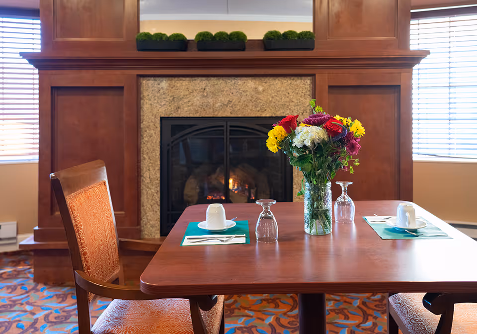 A dining table set for two with cups, glasses, and utensils in front of a lit fireplace. A vase with colorful flowers sits in the center of the table. The room has wooden paneling around the fireplace and windows with blinds on either side.