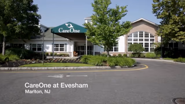 Front exterior view of CareOne at Evesham Assisted Living facility in Marlton, NJ, showing the building entrance with a green canopy labeled 'CareOne', surrounded by trees and landscaping.