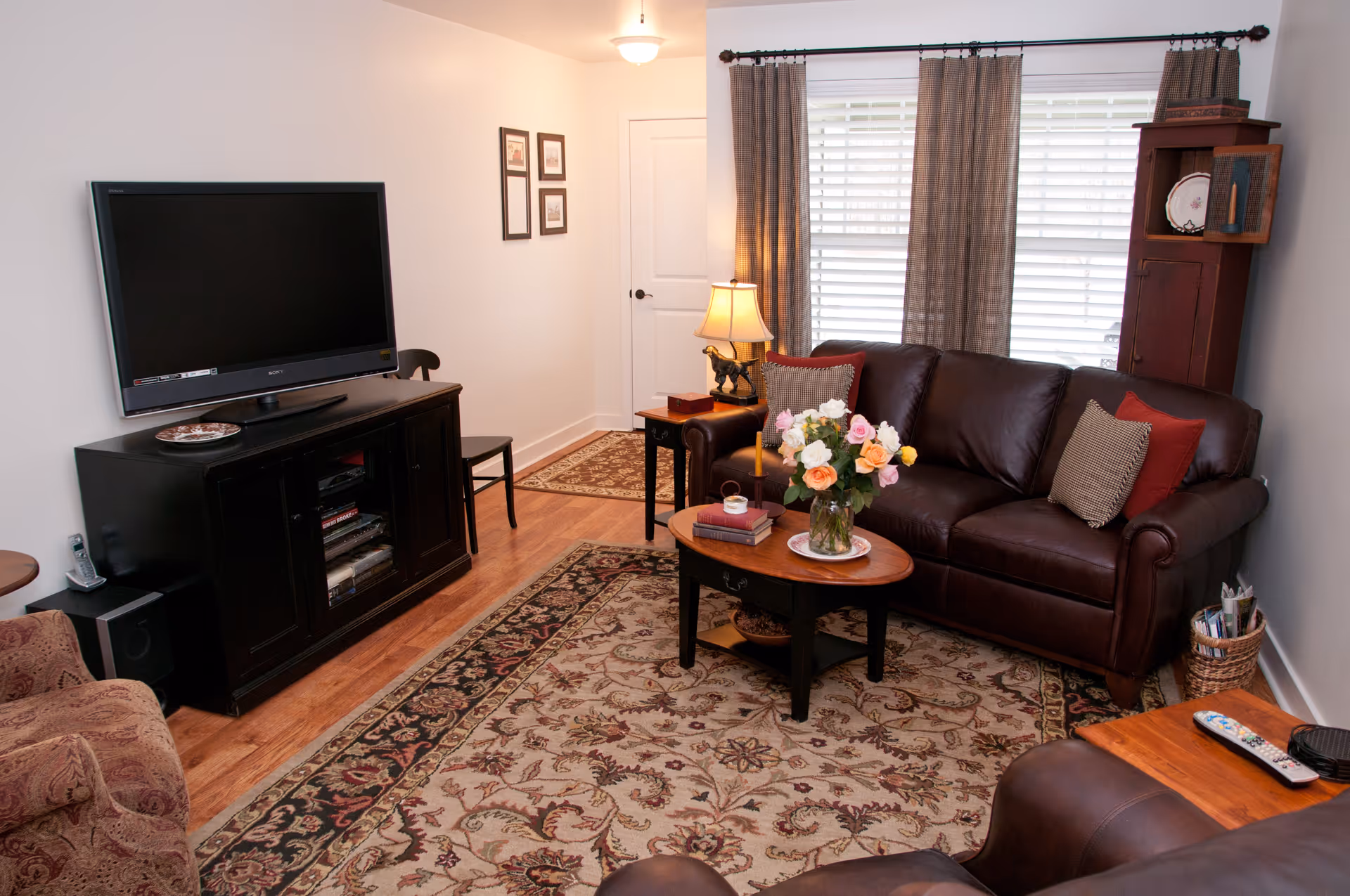 A cozy living room with a dark brown leather sofa adorned with decorative pillows, a wooden coffee table with a vase of colorful flowers, a patterned area rug, a TV on a black stand, a side table with a lamp, and a window with brown curtains.