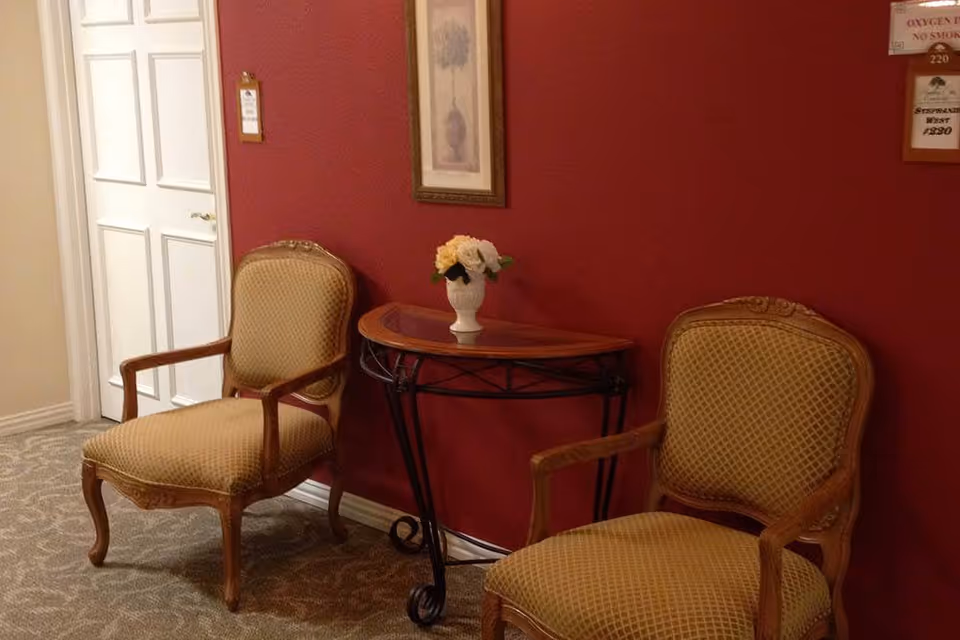 Two upholstered wooden armchairs with patterned fabric are placed on either side of a small half-moon table with a vase of flowers on top, set against a red wall with a framed picture hanging above. A white door is visible to the left, and there are signs on the wall to the right.