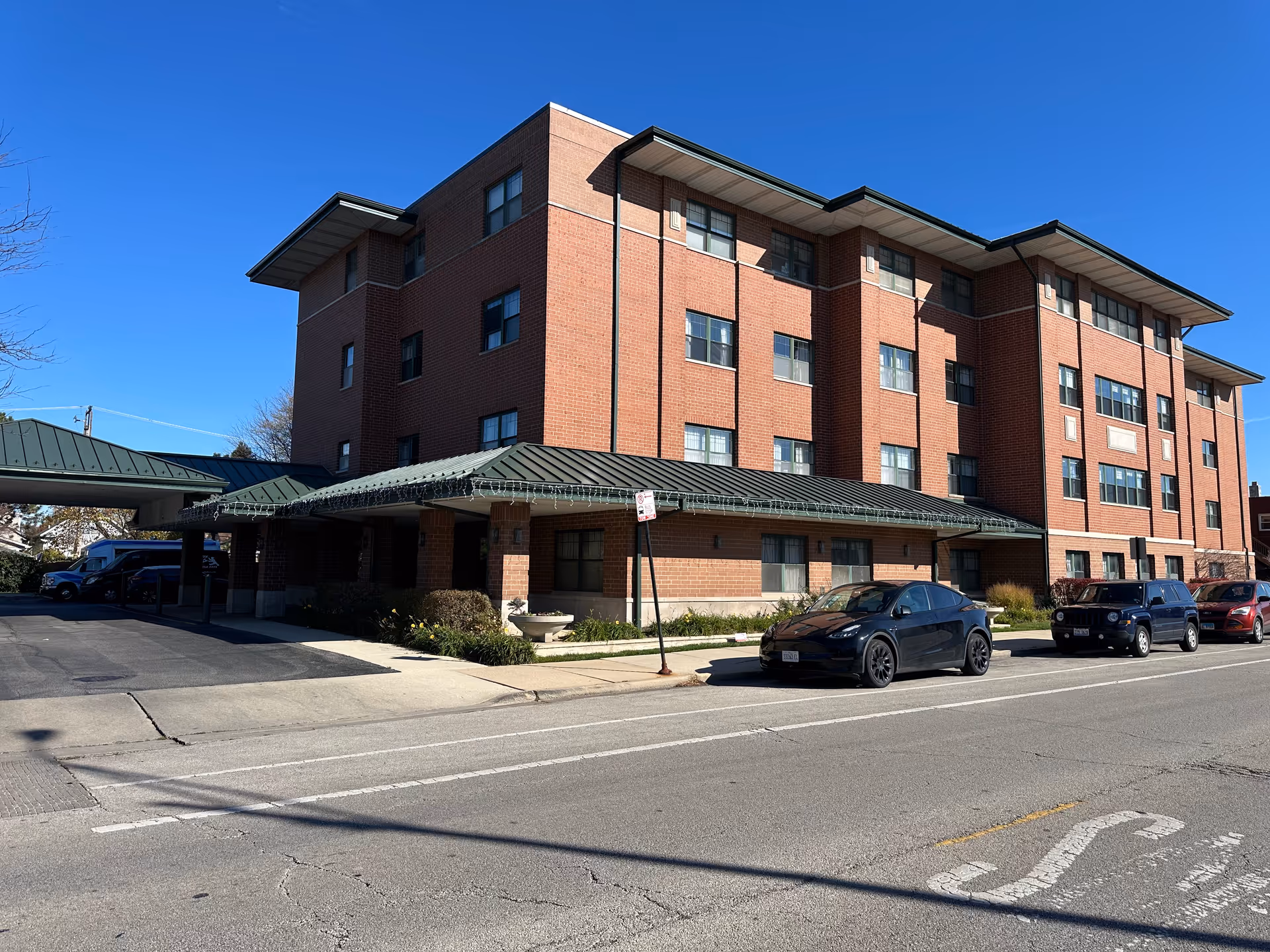 Exterior view of a four-story brick building with a green metal roof canopy over the entrance. Several cars are parked along the street in front of the building under a clear blue sky.