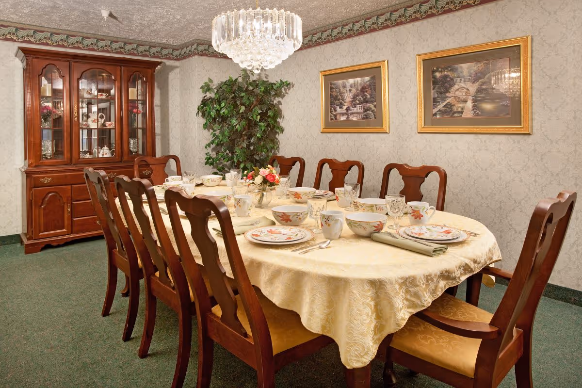 A formal dining room with a long wooden table covered by a cream-colored tablecloth. The table is set with matching floral-patterned dishes, bowls, cups, and glassware. There are eight wooden chairs with cushioned seats around the table. A wooden china cabinet with glass doors displaying decorative items is against the wall. Two framed landscape paintings hang on the wallpapered wall, and a large green potted plant is in the corner. A crystal chandelier hangs above the table.