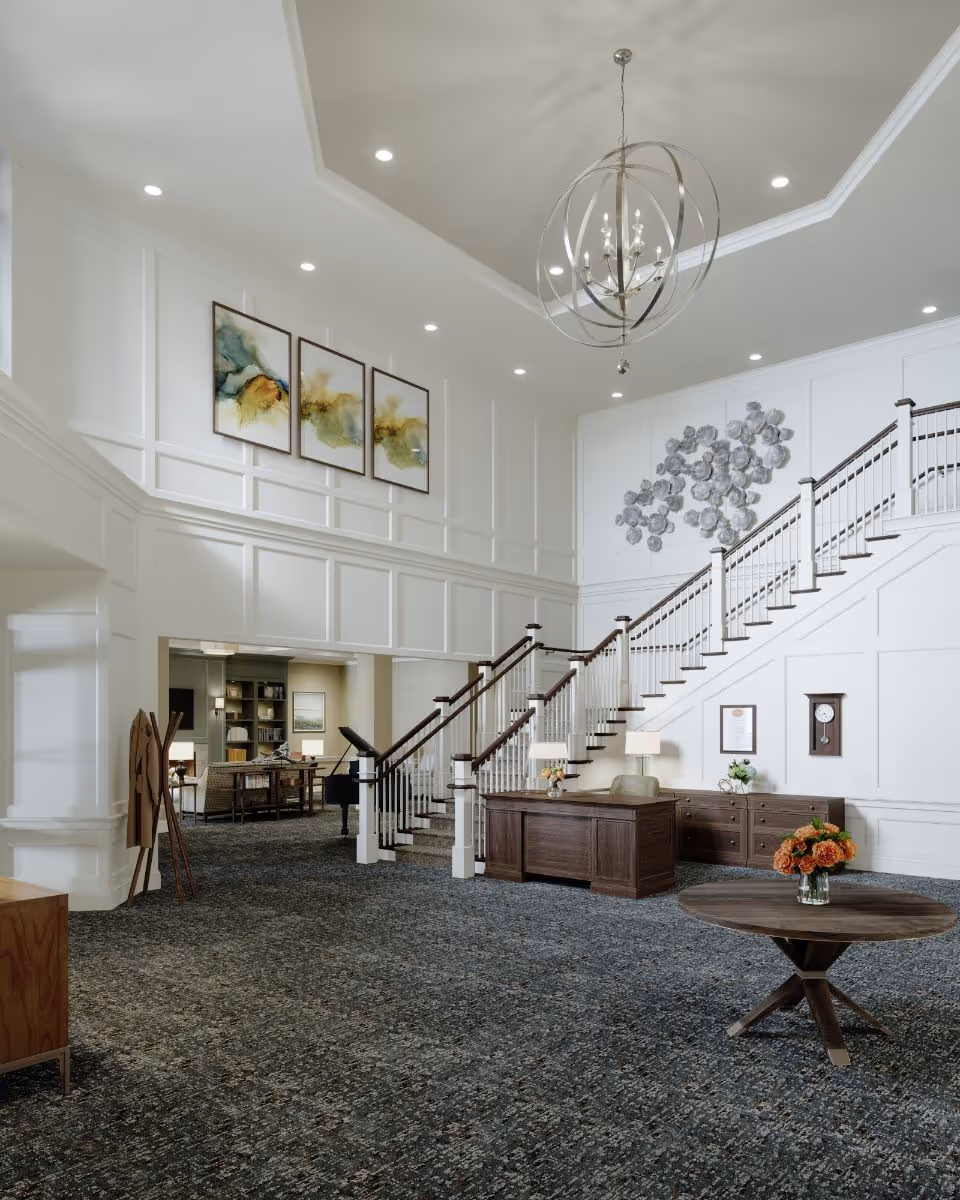 Spacious and elegant interior of a senior living facility with a large staircase, wooden desk, round table with flower vase, and modern chandelier. The walls are white with decorative paneling and artwork, and the carpet is patterned in dark tones.