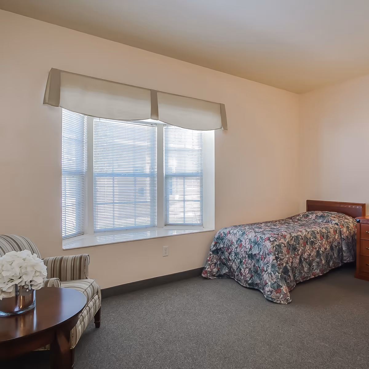 A simple bedroom with a single bed covered in a floral patterned bedspread, a wooden nightstand with drawers, a striped armchair next to a round wooden table with a vase of white flowers, and a large window with blinds and a light-colored valance.