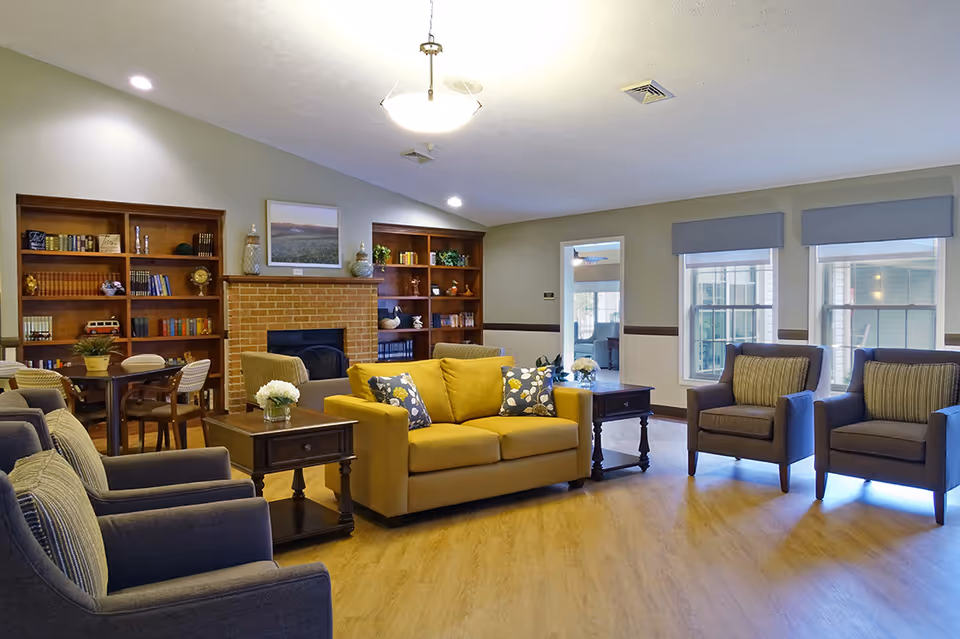 A cozy living room area in Colonial Courtyard at Bedford featuring a yellow sofa with patterned cushions, two gray armchairs with striped pillows, wooden side tables with flower vases, a brick fireplace flanked by built-in wooden bookshelves filled with books and decorative items, and large windows with gray valances letting in natural light.