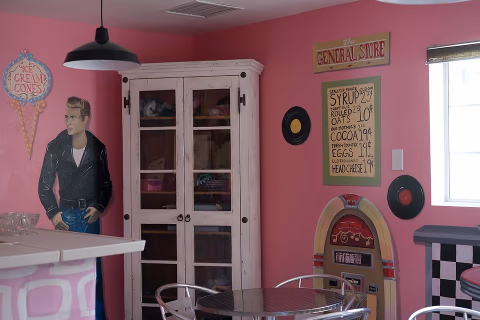 A retro-themed room with pink walls featuring a painted figure of a man in a leather jacket, a white cabinet with glass doors, a round metal table with chairs, and wall decorations including a sign that reads 'The General Store', a menu board with prices for syrup, oats, cocoa, eggs, and head cheese, two vinyl records, and a jukebox illustration. A window with a bamboo shade lets in natural light.