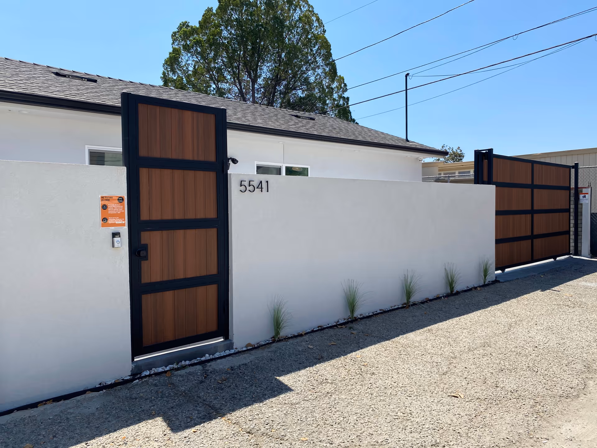 Exterior view of a white wall with a wooden pedestrian gate and a matching larger wooden gate, both framed in black metal. The wall has the number 5541 displayed on it, with small plants growing along the base. A house with a gray roof and windows is visible behind the wall under a clear blue sky.