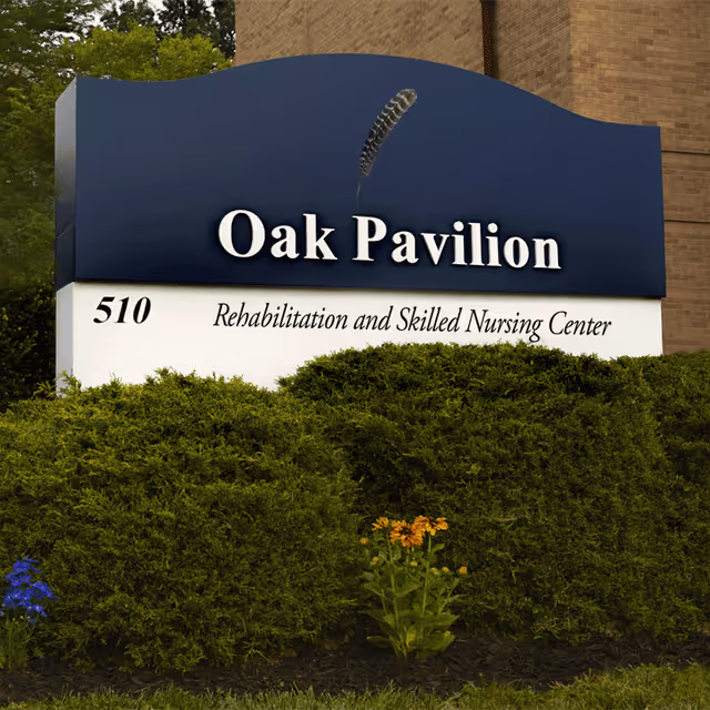 Outdoor sign for Oak Pavilion Rehabilitation and Skilled Nursing Center with the number 510, surrounded by green bushes and some flowers in front of a brick building.