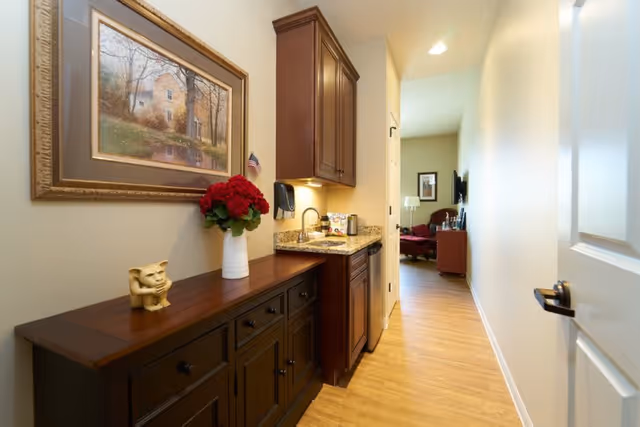 A narrow hallway with wooden flooring leading to a living area. On the left side of the hallway, there is a dark wooden cabinet with a small decorative sculpture and a vase of red flowers. Above the cabinet hangs a framed painting of a house in a wooded area. Next to the cabinet is a small kitchenette with a granite countertop, a sink, a coffee maker, and upper wooden cabinets. The living area at the end of the hallway has a red armchair, a floor lamp, and a wall-mounted TV.