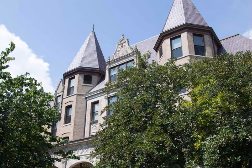 Exterior view of a large, historic-style building with pointed towers and multiple windows, partially obscured by green trees under a blue sky with some clouds.