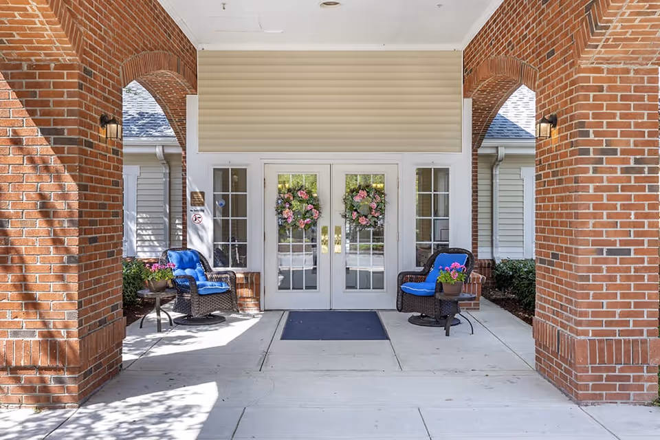 Entrance of a building with double glass doors decorated with floral wreaths. The entrance is framed by red brick arches on both sides. Two wicker chairs with blue cushions and small tables with potted flowers are placed on either side of the doors. The area is well-lit with wall-mounted lights on the brick columns.