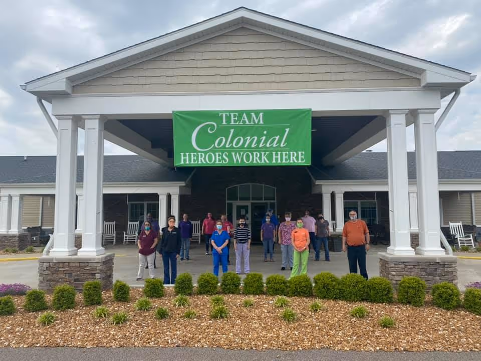 A group of staff members standing outside the entrance of Colonial Assisted Living & Independent Living facility under a large green banner that reads 'TEAM Colonial HEROES WORK HERE'. The building has a covered entrance with white pillars and stone bases, and there are bushes and mulch in the foreground.