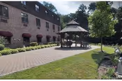 Outdoor paved courtyard area with a gazebo in the center, surrounded by a well-maintained lawn and landscaping. A brick building with multiple windows and red awnings is visible on the left side, and trees are in the background under a partly cloudy sky.
