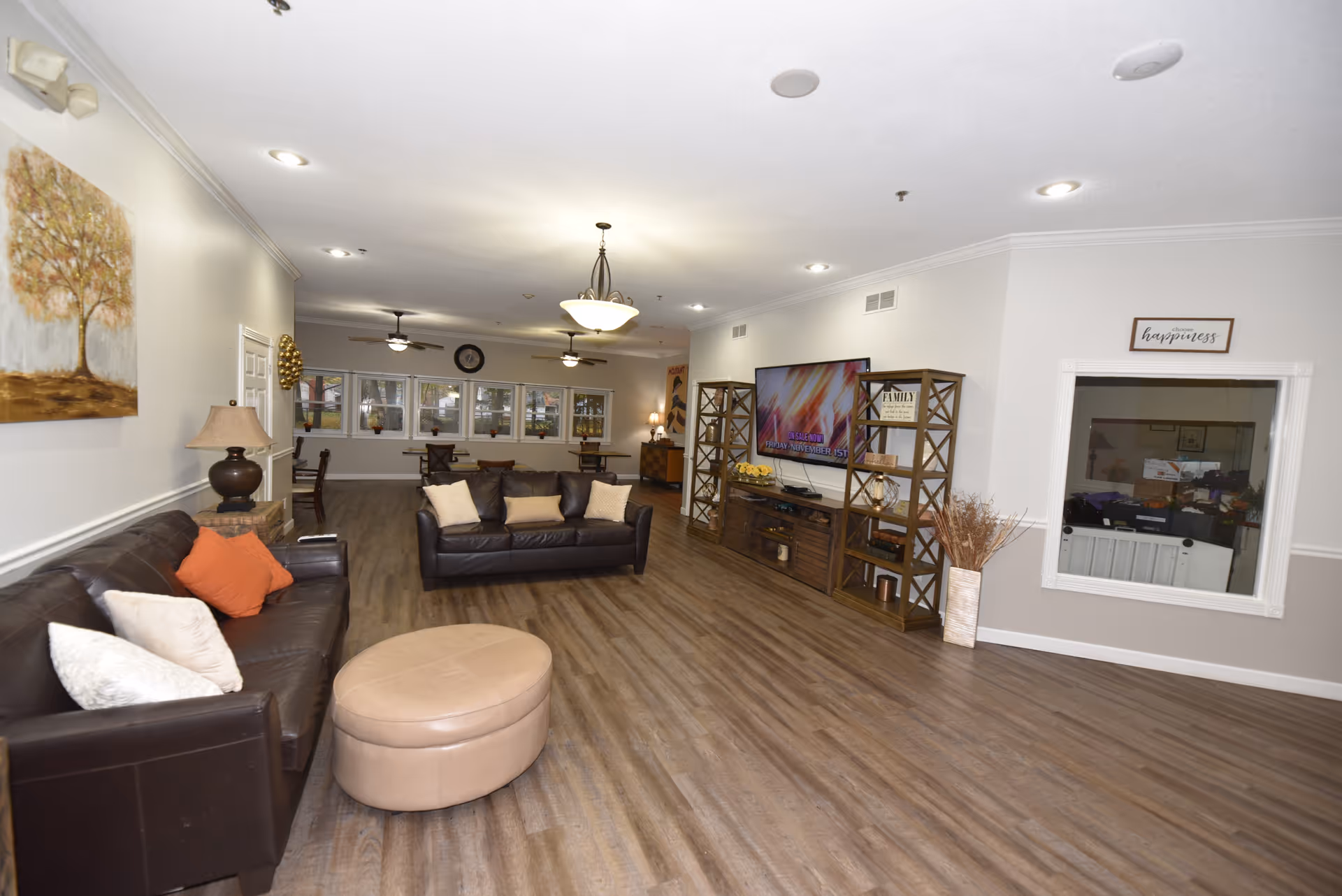 A spacious living room area with two dark brown leather sofas, a round beige ottoman, and a large flat-screen TV mounted on the wall. The room has wood flooring, light-colored walls, ceiling lights, and ceiling fans. There is a painting of a tree on one wall and a window with a sign above it that reads 'Choose happiness'. In the background, there are tables and chairs near windows.