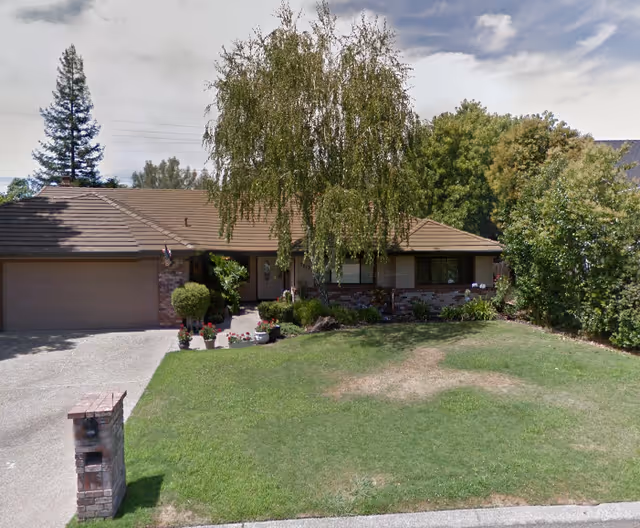 Single-story residential building with a brown tiled roof, brick and wood exterior, surrounded by green trees and shrubs. There is a driveway leading to a two-car garage and a grassy front yard with some patches of dry grass. A mailbox is visible near the street.