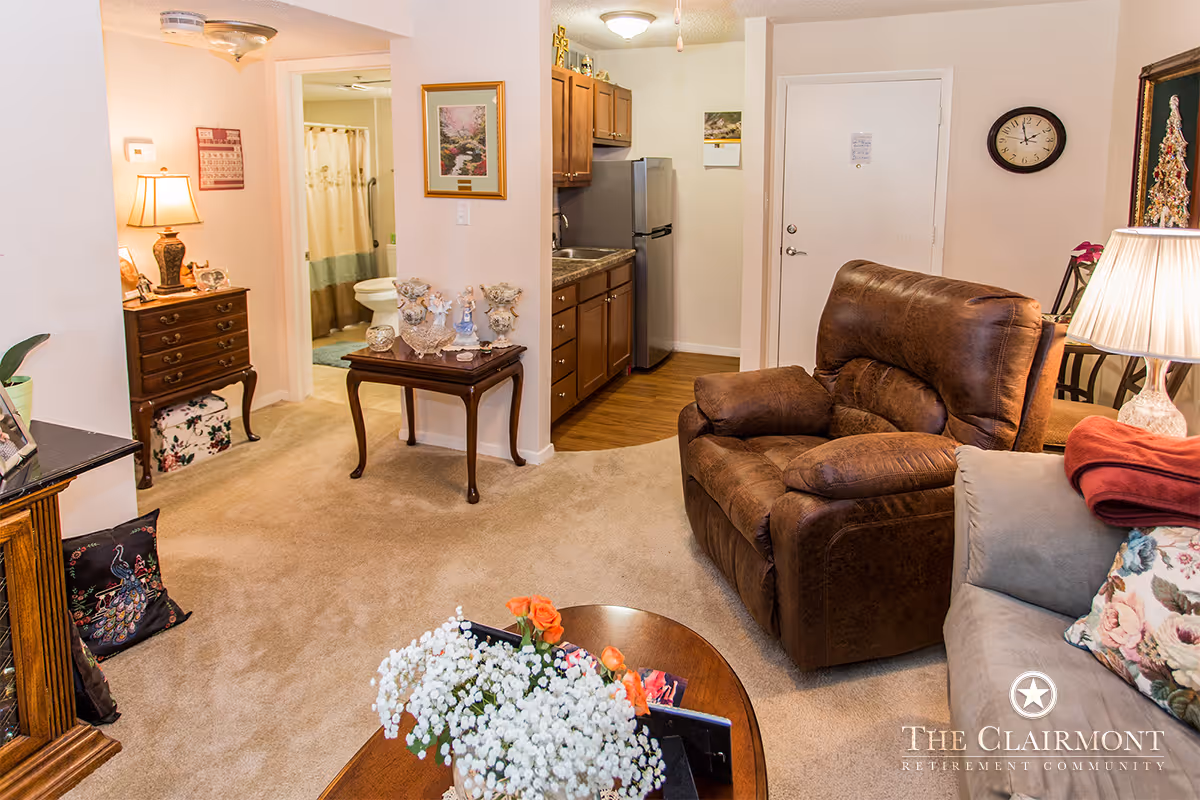 A cozy living room area with a brown leather recliner and a gray sofa with floral pillows. A wooden coffee table with a flower arrangement is in the foreground. To the left, there is a small wooden table with decorative items and a lamp on a chest of drawers. The room opens into a small kitchen area with wooden cabinets and a stainless steel refrigerator. A bathroom with a shower curtain is visible through an open door. The walls are light-colored, and the floor is carpeted except for the kitchen area which has wooden flooring.
