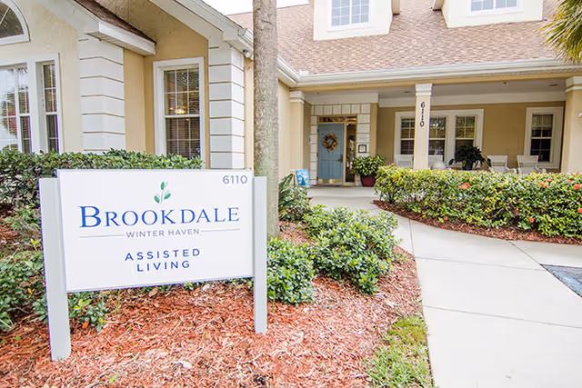 Exterior view of the entrance to Brookdale Winter Haven assisted living facility with a white sign displaying the facility name and address number 6110, surrounded by landscaping and a walkway leading to the front door.