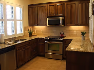 A kitchen with wooden cabinets, granite countertops, a stainless steel oven with a red kettle on top, a microwave above the oven, a double sink under a window with white shutters, and potted plants on the countertops.