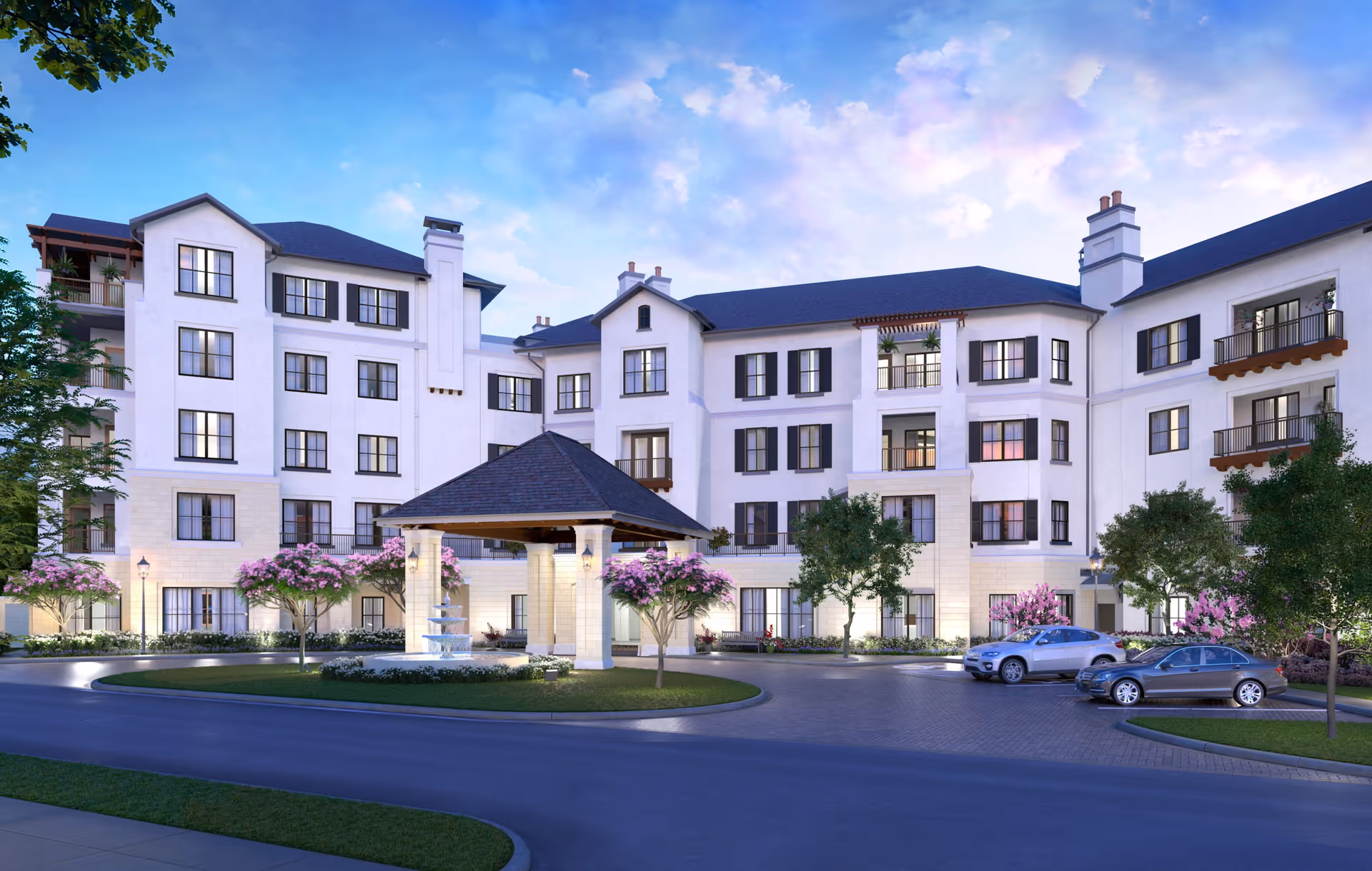 Exterior view of a multi-story senior living facility building with white walls, black window shutters, and a covered entrance with a fountain in front. There are trees and flowering plants around the driveway, and a few cars parked nearby under a partly cloudy sky at dusk.