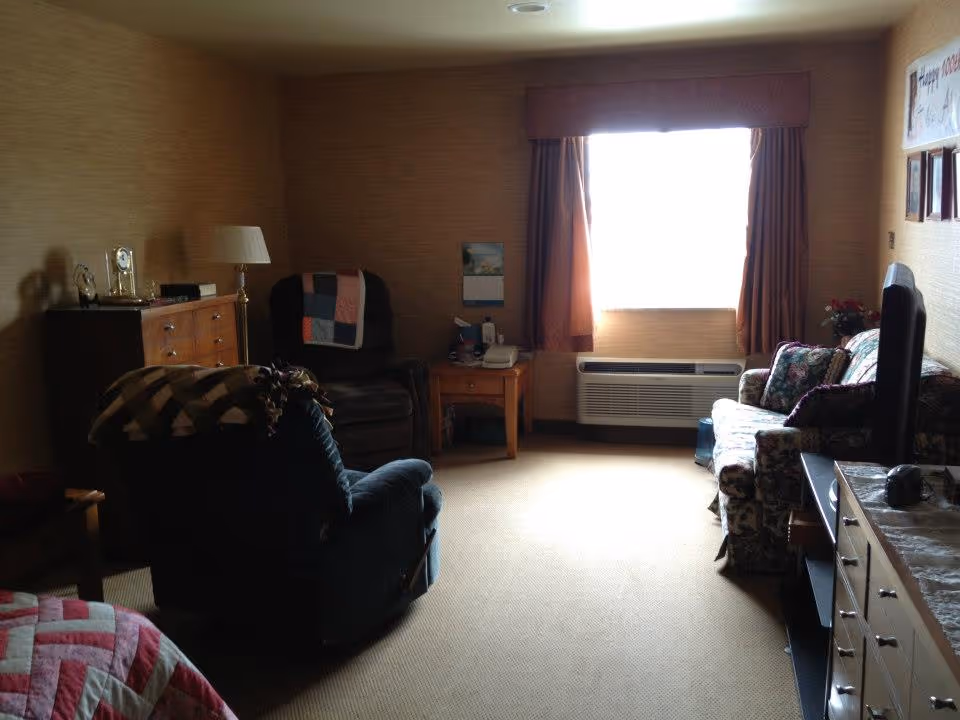 A cozy living room in an assisted living facility with a window covered by curtains letting in bright light. The room contains a floral-patterned sofa, two armchairs with blankets draped over them, a wooden dresser with a lamp and decorative items, a side table with a telephone, and a television on a stand.