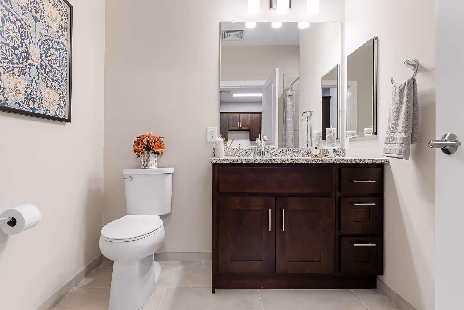 A clean and modern bathroom featuring a white toilet with a vase of orange flowers on top, a dark wood vanity with a granite countertop, a large mirror above the sink, a towel hanging on a hook on the wall, and a framed floral artwork on the left wall.