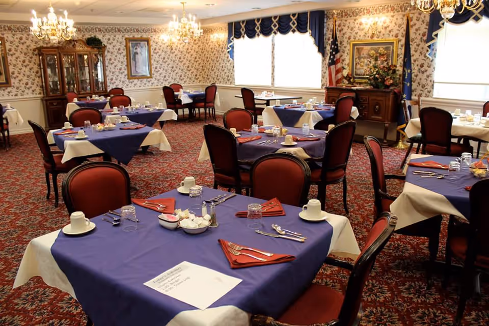 A dining room with multiple tables covered in blue and white tablecloths, set with cups, glasses, silverware, and red napkins. The room has floral wallpaper, chandeliers, and red upholstered chairs. There is a wooden cabinet with glass doors and framed artwork on the walls.