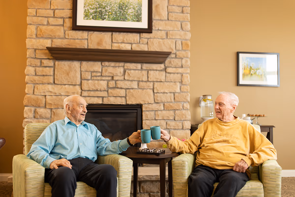 Two elderly men sitting in armchairs in a cozy room with a stone fireplace behind them. They are smiling and clinking blue mugs together in a toast. The room has warm beige walls with framed pictures and a small table between the chairs holding a chessboard and other items.