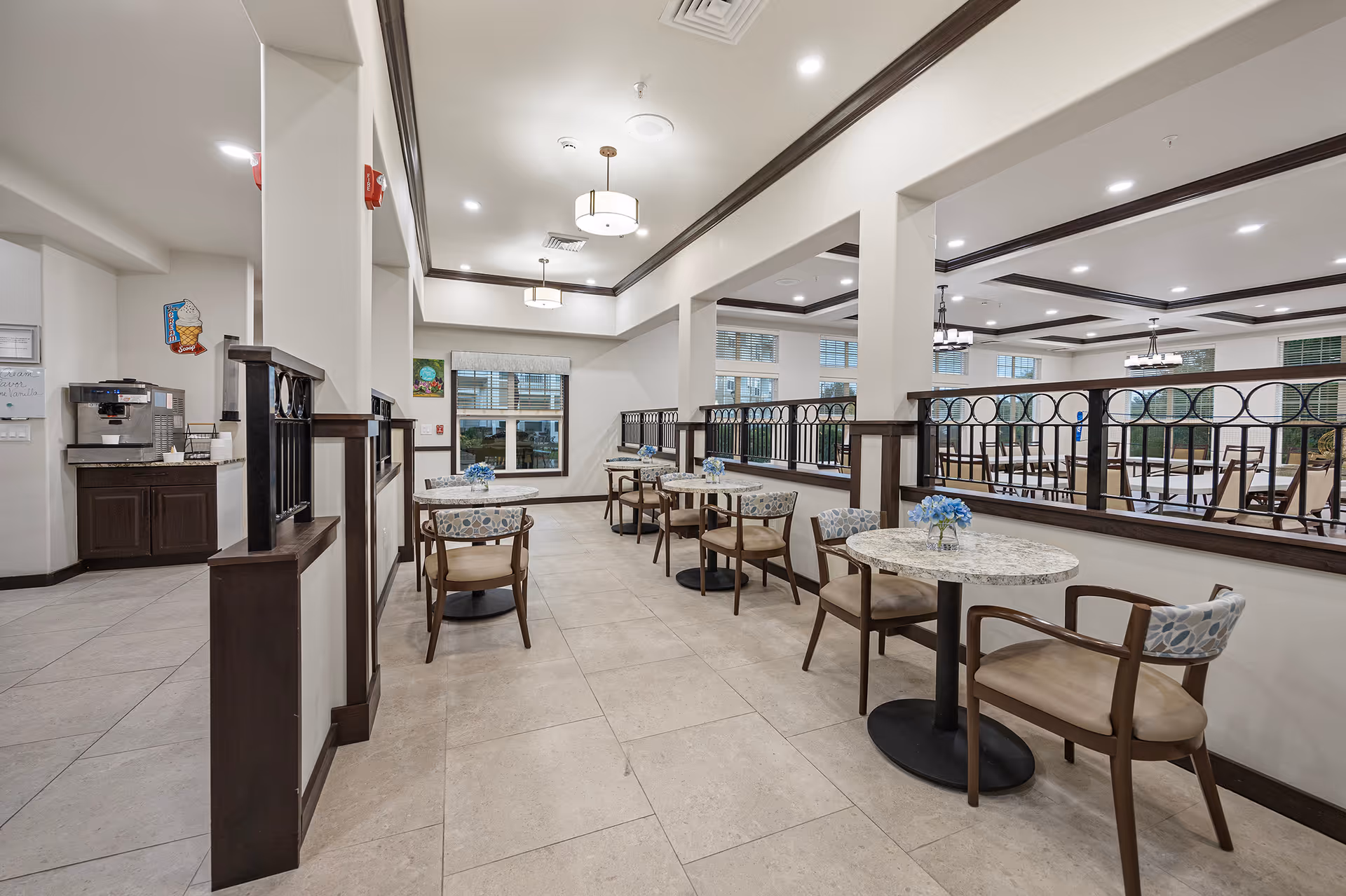 Interior view of a dining area in a retirement living facility with small round tables and chairs arranged neatly. The space is well-lit with ceiling lights and has a coffee station on the left side. The area features light-colored walls, tiled floors, and dark wood trim accents.