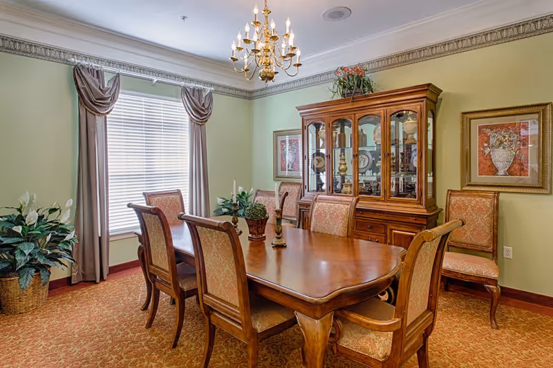 A formal dining room with a wooden dining table surrounded by eight upholstered chairs. The room features a large window with curtains, a chandelier hanging from the ceiling, a wooden china cabinet displaying decorative items, framed artwork on the walls, and potted plants in the corners.