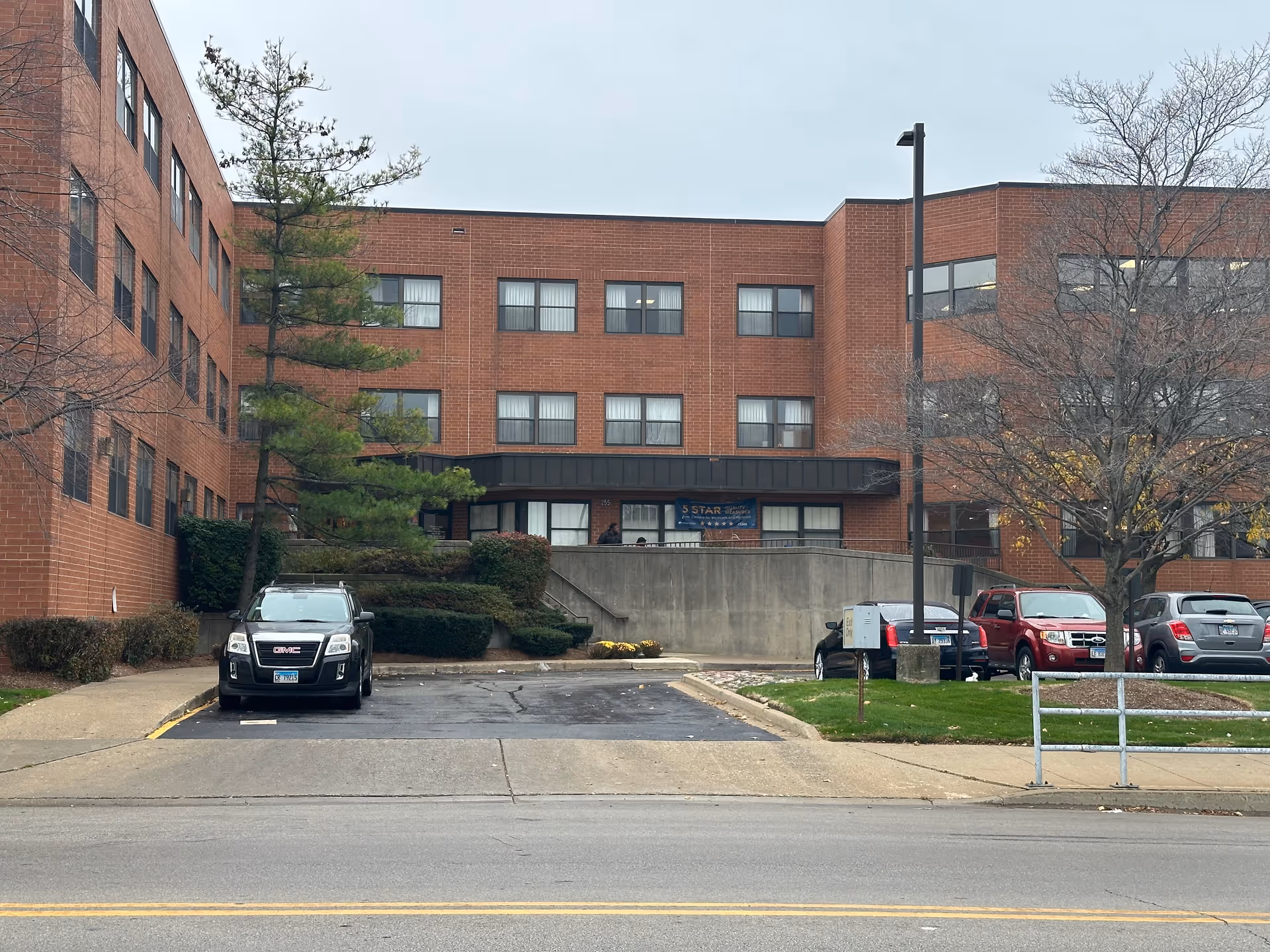 Exterior view of a multi-story brick building with several windows, a small parking area with cars, a tree with no leaves, and a banner on the building reading '5 STAR'. The scene is overcast with a gray sky.