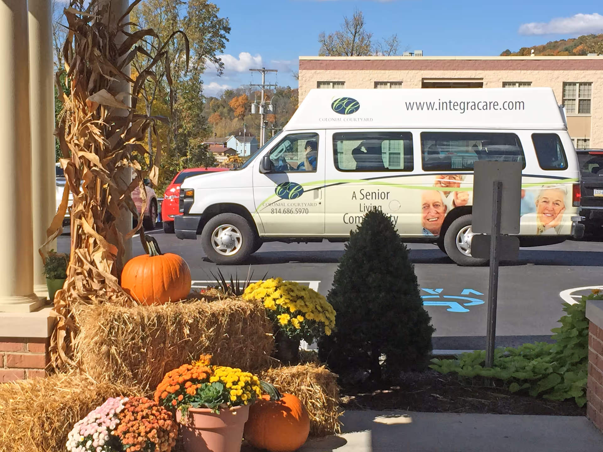 View of a parking lot outside a senior living facility with a white van parked. The van has the Colonial Courtyard logo and website www.integracare.com on it. In the foreground, there are autumn decorations including pumpkins, hay bales, and colorful potted flowers near a brick column and a small evergreen shrub.