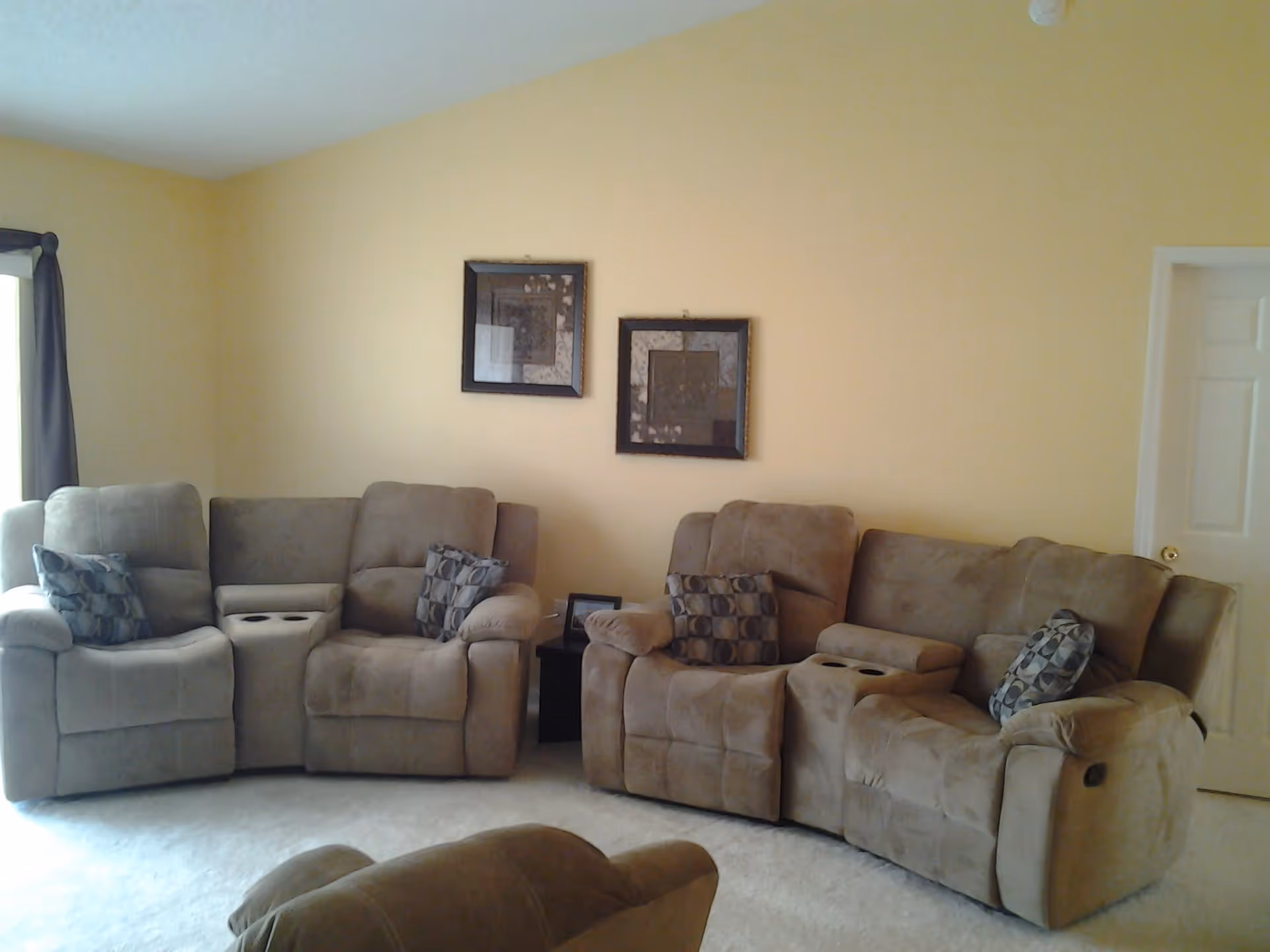 A living room with beige walls and carpet, featuring two brown reclining sofas with patterned cushions, a small dark wooden side table between them, and two framed artworks hanging on the wall above the sofas. A window with dark curtains is partially visible on the left side, and a white door is on the right side of the room.