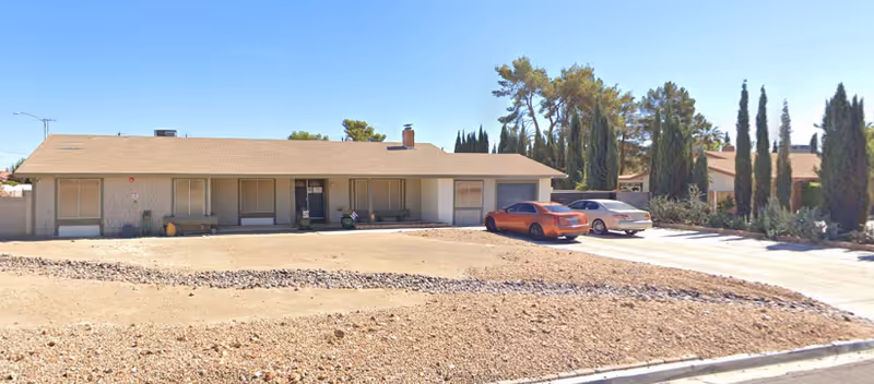 Single-story residential building with a beige exterior and a brown shingle roof. There are several windows with closed blinds and a central entrance door. Two cars are parked on the driveway to the right of the building. The front yard is mostly bare dirt with a strip of rocks running through it. Tall trees and shrubs are visible in the background under a clear blue sky.