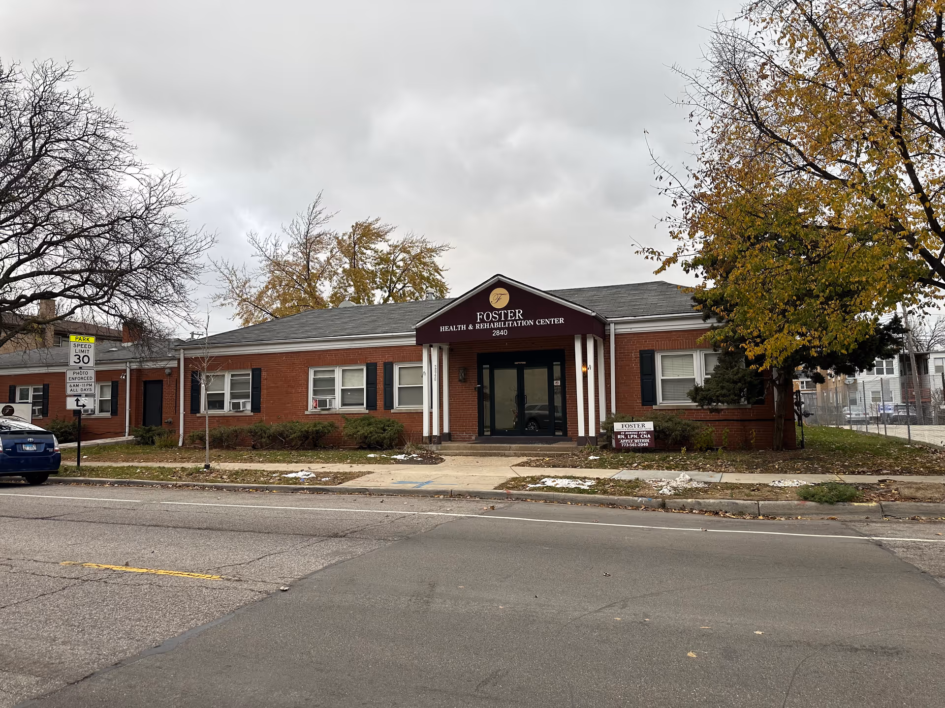 Exterior view of Foster Health & Rehabilitation Center, a single-story brick building with a central entrance covered by a maroon awning displaying the facility's name and address. There are trees with autumn foliage on either side of the building and a street with a speed limit sign in front.
