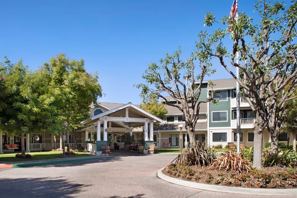 Exterior view of Ivy Park at Salinas senior living facility showing a driveway with a covered entrance, landscaped garden with trees and plants, and a multi-story building with balconies and windows under a clear blue sky.