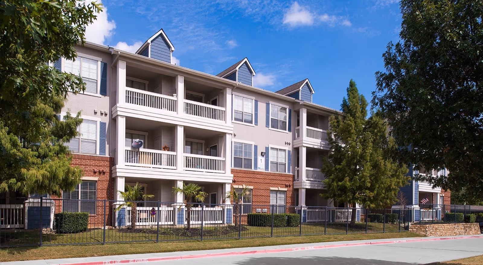 Exterior view of a three-story residential building with balconies, surrounded by trees and a fenced lawn under a blue sky with some clouds.