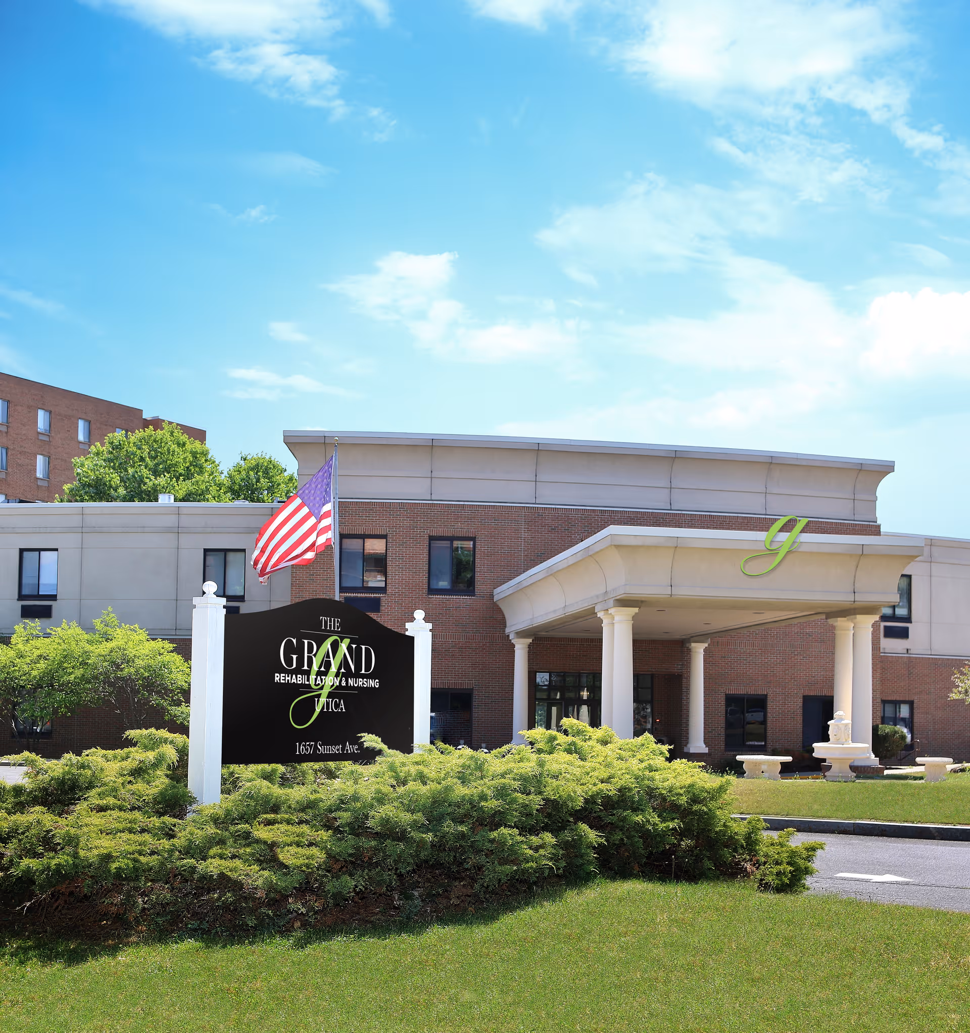Exterior view of The Grand Rehabilitation and Nursing at Utica building with a large entrance canopy supported by columns, an American flag flying on a pole, and a black sign with the facility's name and address surrounded by green bushes and grass under a blue sky with some clouds.